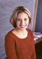 Professional headshot of a woman with blonde hair wearing an orange-red sweater against a neutral background.