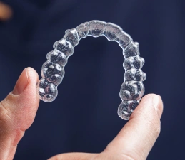 Clear plastic orthodontic retainer or aligner held between two fingers against dark background.