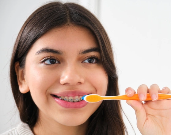 Young woman brushing teeth with orange toothbrush, smiling at camera in bright setting.