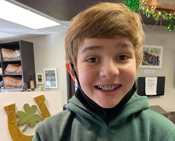 Young boy smiling at camera in orthodontics office with decorative posters on wall behind him.