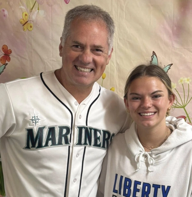 Two people wearing baseball jerseys smile together in front of a decorative floral backdrop.