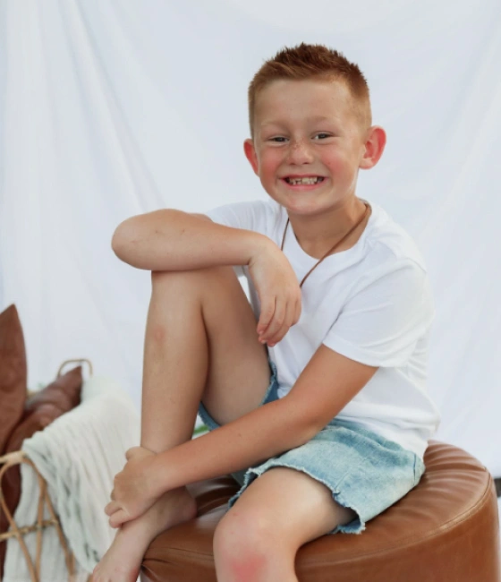 Smiling young boy sitting on brown chair wearing white shirt against white background.