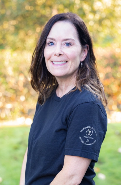 Woman wearing navy Tingey Orthodontics polo shirt smiling outdoors with blurred green background.