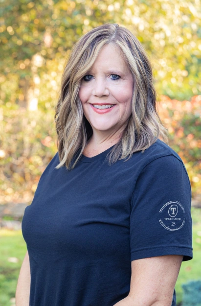 Woman in navy blue Tingey Orthodontics shirt smiling outdoors with blurred green background.