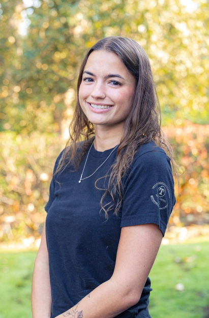 Young woman smiling at camera wearing dark blue shirt, outdoor autumn setting with blurred green foliage background.
