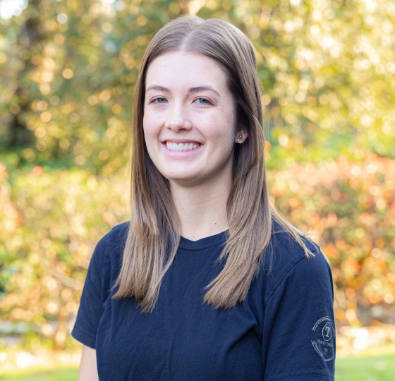 Young woman smiling at camera wearing dark blue shirt, outdoors with blurred golden foliage background.