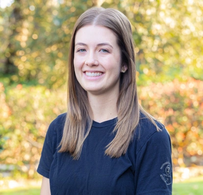 Woman smiling at camera wearing navy blue shirt, autumn foliage background, professional headshot.
