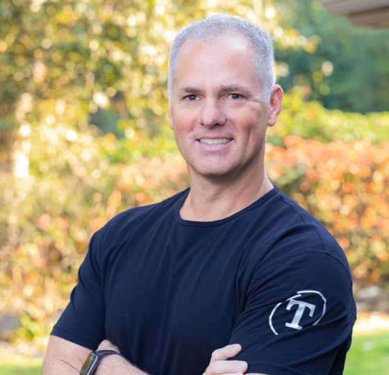 Professional male orthodontist posing outdoors with arms crossed, wearing branded black t-shirt against blurred garden background.