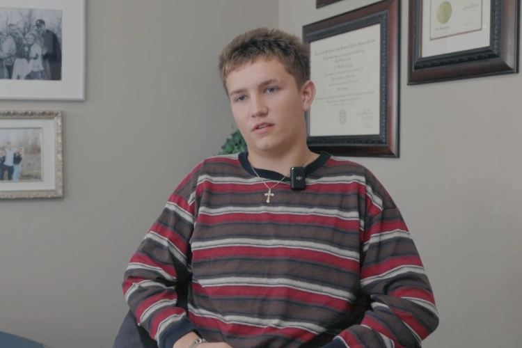 Young man in striped rugby shirt sitting in orthodontic office with framed certificates on wall.