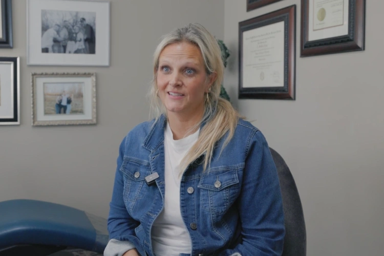 Woman in denim jacket smiling in orthodontic office with framed certificates on wall behind her.