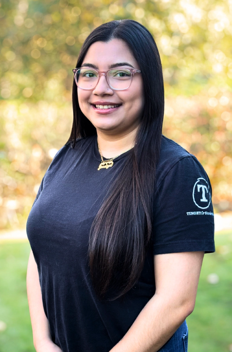 Young woman wearing glasses and black Tingey Orthodontics shirt smiling outdoors with blurred background.