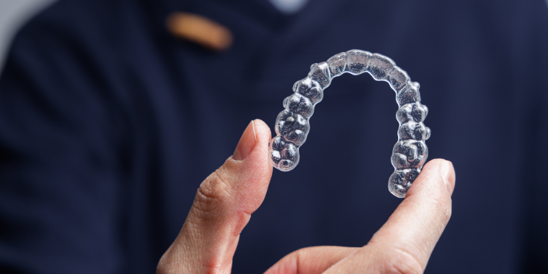 Person holding a clear Invisalign aligner tray between their fingers against a dark background.
